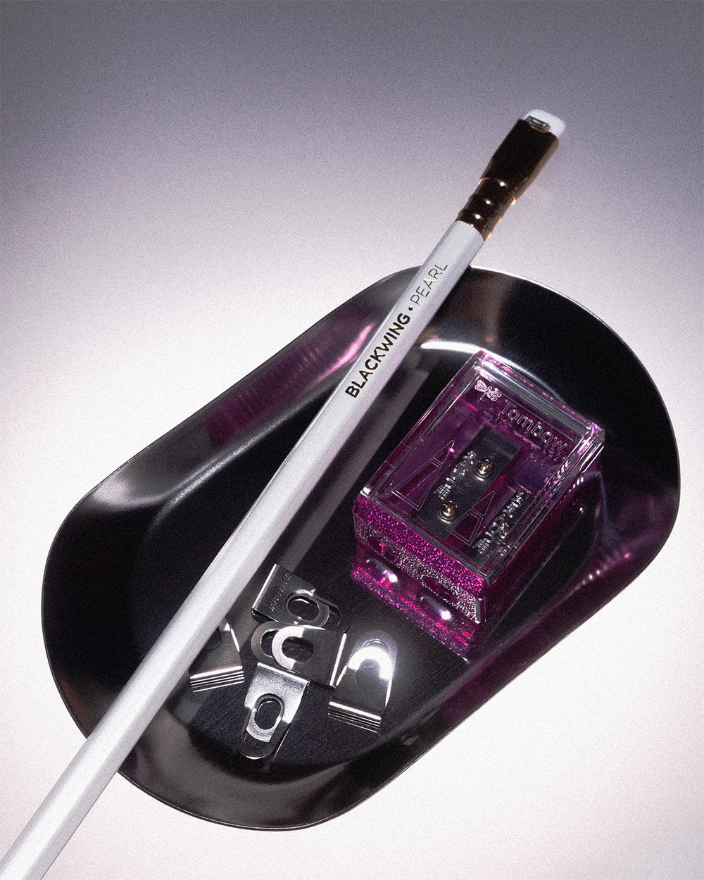 A silver tray on a gradient background. In oval silver tray holds a Blackwing Pearl wooden pencil, some Super Clips from OHTO, and a pink translucent pencil sharpener from Japanese brand, Tombow.