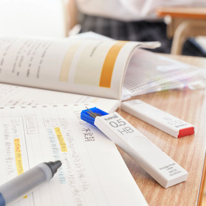 A close-up of a desk displays an open notebook with highlighted notes, a gray pen, and two Uni Smudge-proof Lead Refills (0.5 HB). A book and folders can be seen in the background.