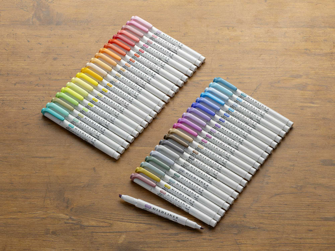 Two Zebra Mildliner - Single Marker highlighters are neatly arranged diagonally on a wooden surface, with a purple Mildliner lying horizontally below them.