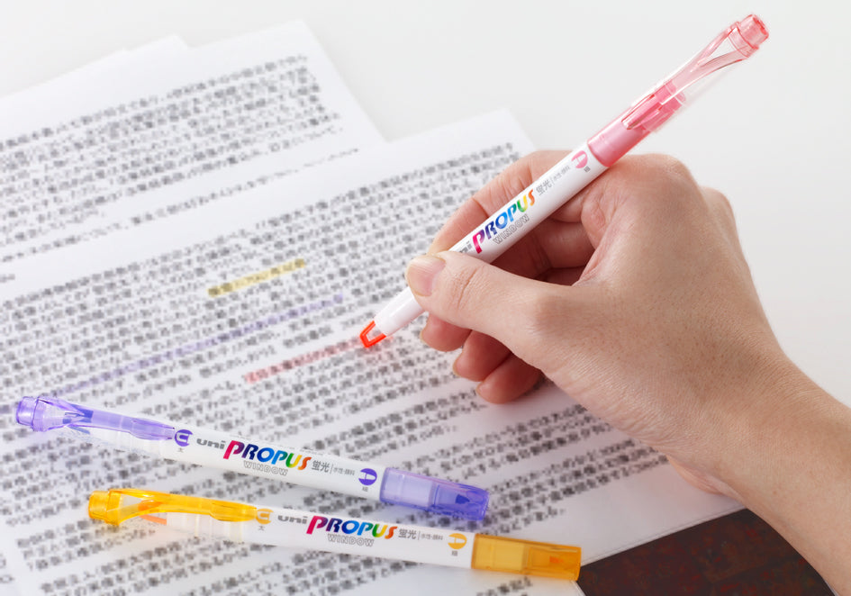 Hand holding a pink Propus highlighter over printed text with additional highlighters on a white surface.
