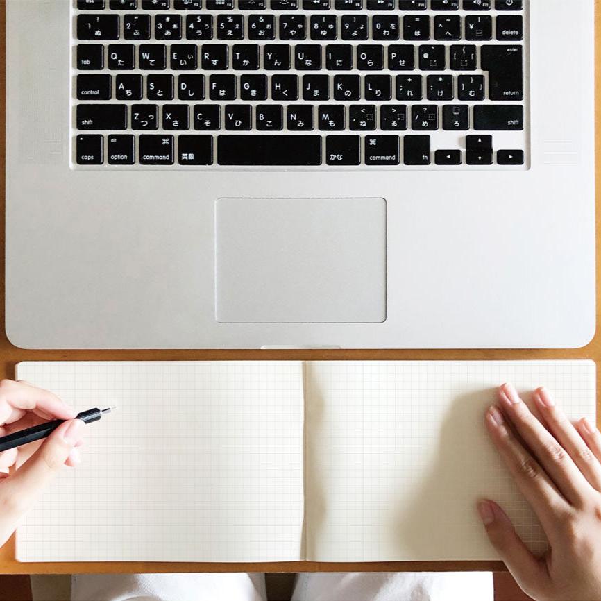 A person holds a pen above an open Isshoni Laptop Notebook - Grid, placed on a desk beneath a 13 laptop keyboard.