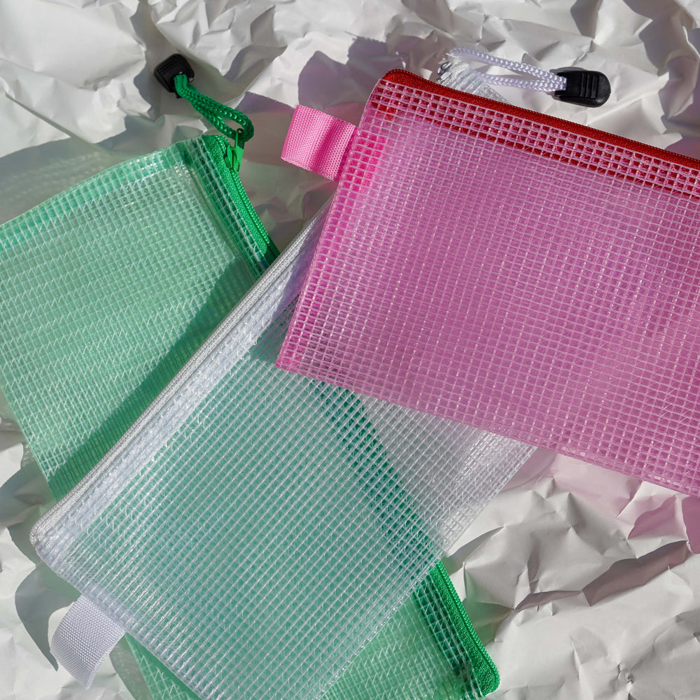 Three TACTO STUDIO Mesh Cases in green, white, and pink with zip closures overlap on crumpled white paper, sunlight casting shadows through each transparent pouch and highlighting the textured surface.