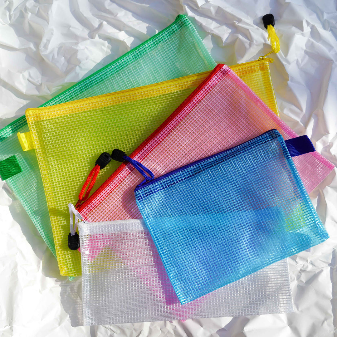 Five TACTO STUDIO Mesh Cases in green, yellow, pink, blue, and white are stacked on a crumpled white surface. Each PVC pouch is transparent and features a sturdy zip closure with a small black zipper pull.
