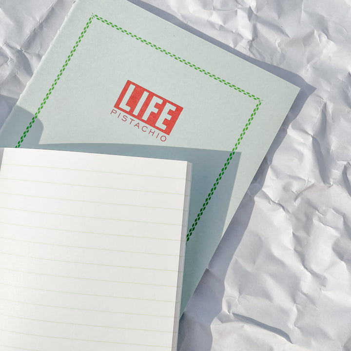 A closed LIFE Pistachio Notebook with PISTACHIO on the cover rests on wrinkled white paper. An open, fountain pen-friendly lined LIFE Pistachio Notebook partially covers the lower part of the closed one.