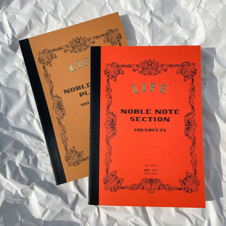 Two LIFE Noble Notebook - 100 Sheets, made in Japan, one with a brown Plain cover and one with an orange Section cover, rest on crumpled white paper. Both have black spines, decorative borders, and fountain pen-friendly paper.