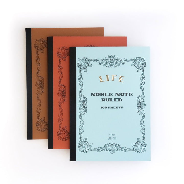 Three LIFE Noble Notebooks, made in Japan, are stacked in light blue, orange, and brown covers with ornate black borders and “Noble Notebook - 100 Sheets” branding. The paper is fountain pen-friendly.