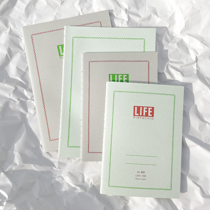 Four light green LIFE Pistachio Notebooks with decorative red and green borders are displayed on crumpled white paper under bright lighting—fountain pen-friendly and made in Japan.