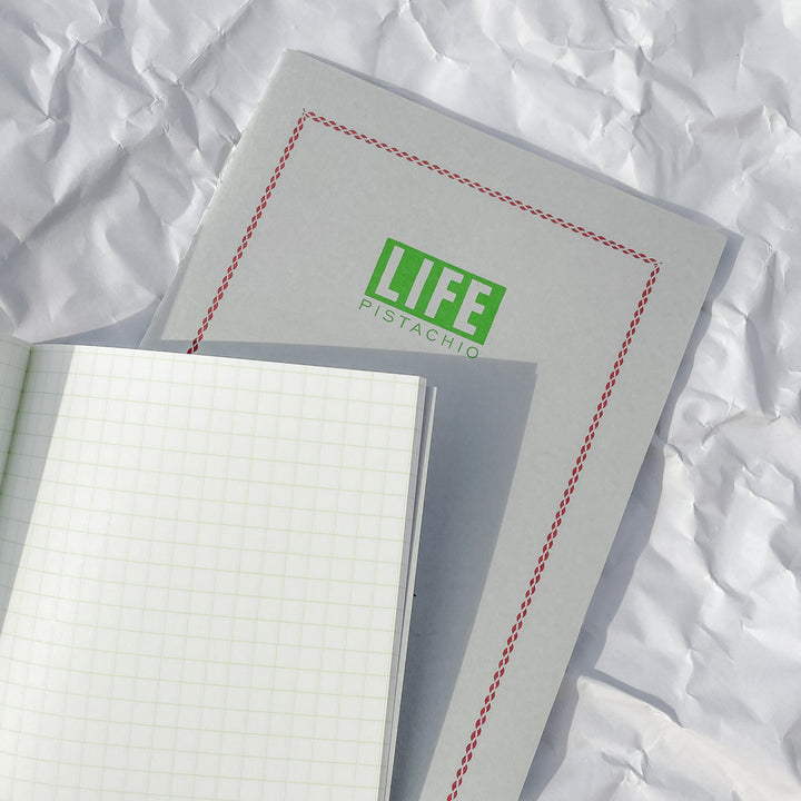 A closed LIFE Pistachio Notebook with PISTACHIO on the cover rests on wrinkled white paper. An open, fountain pen-friendly lined LIFE Pistachio Notebook partially covers the lower part of the closed one.