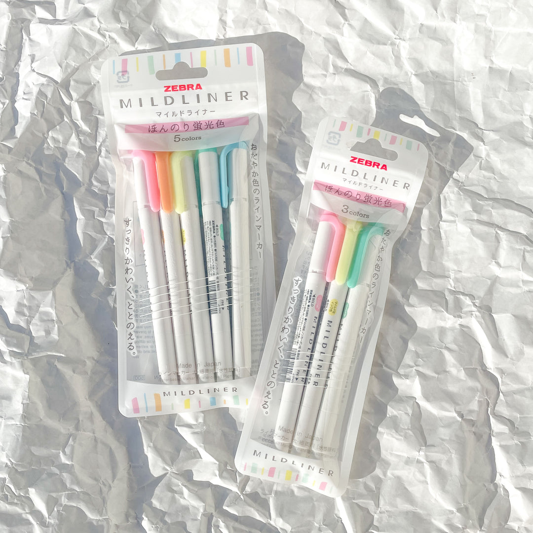Two packs of Zebra Mildliner Highlighter Markers—Set of 3 and Set of 5, Light Fluorescent—rest on crumpled white paper. Each marker has a white body with pastel caps, features water-based ink, and a dual-tip design.