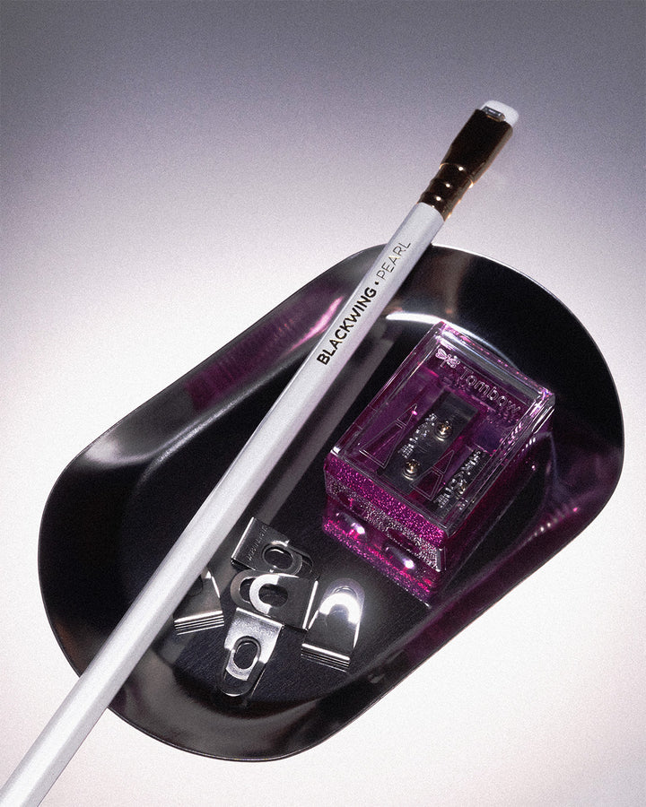 A silver tray on a gradient background. In oval silver tray holds a Blackwing Pearl wooden pencil, some Super Clips from OHTO, and a pink translucent pencil sharpener from Japanese brand, Tombow. 
