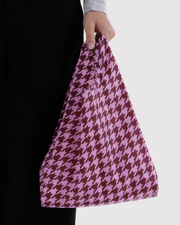 A person holding a large houndstooth pattern tote bag in pink and dark brown, shown against a light gray background.