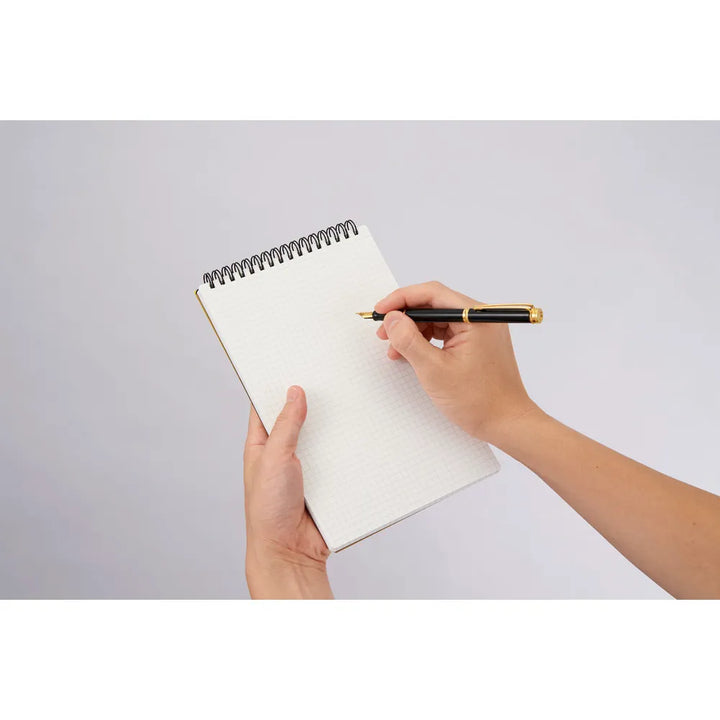 Hand holding a pen poised over an open notebook on a light gray  #paper-type_5mm-grid